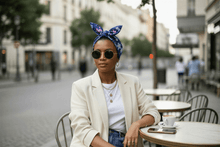 Load image into Gallery viewer, Blue floral cotton hair scarf tied in a bow, worn by a woman with vintage sunglasses, seated at an outdoor café.