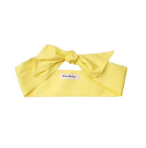 A yellow cotton hair scarf laid out on a white background, showing a bow knot at one end with a tag visible.