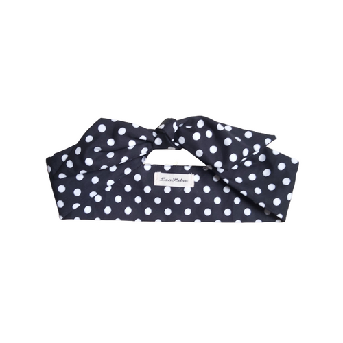 A black polka dot cotton hair scarf displayed on a hanger against a textured background.